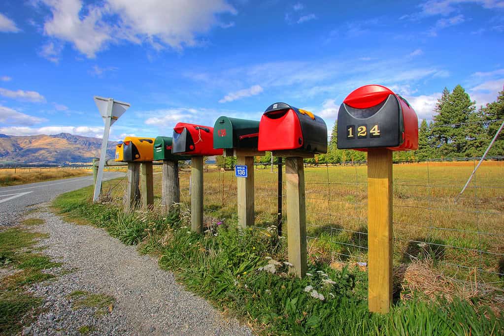 Colorful mailboxes on wooden posts along a rural roadside with scenic mountain and blue sky backdrop.